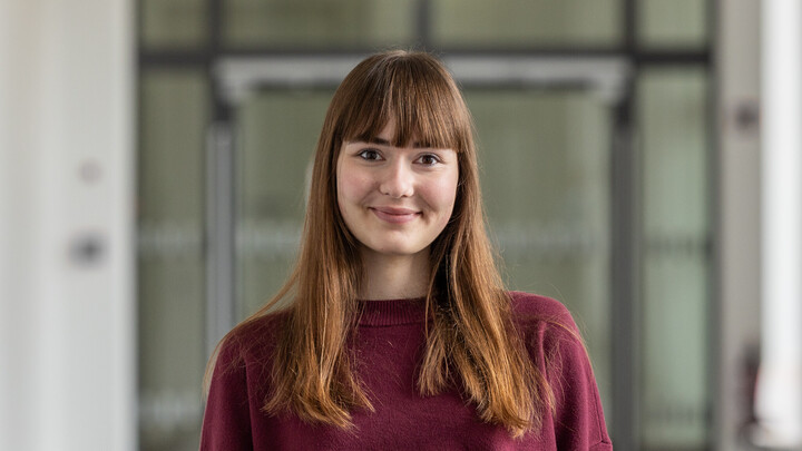 Portrait of Anna Aigner. She's wearing a maroon coloured jumper standing in front of a glass door