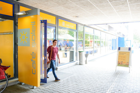 Ein Student verlässt den blau gelben Eingang des "Café Soleil" der Universität Mannheim in A5.