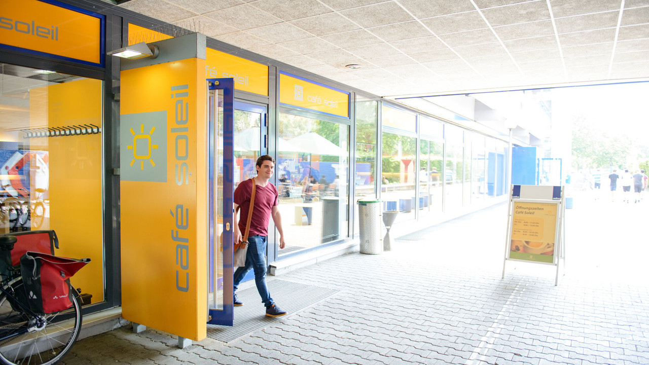 Ein Student verlässt den blau gelben Eingang des "Café Soleil" der Universität Mannheim in A5.