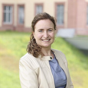Imola Czolbe hat lockige, zu einem Pferdeschwanz gebundene Haare. Sie trägt Creolen-Ohrringe und eine helle Lederjacke. 