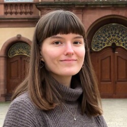 Portät von Luisa Gehm. Luisa Gehm trägt einen dunklen Rollkragenpulli und dezenten Goldschmuck. Sie hat braune Augen und braune Haare, die ihr bis über die Schulter reichen. Zudem trägt sie einen akkuraten Pony-Schnitt. 