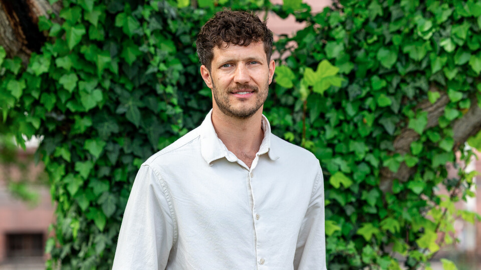 Porträt von Prof. Olbert. Er steht vor einer begrünten Wand, trägt ein weißes Hemd und lächelt in die Kamera.