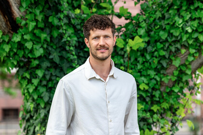 Porträt von Prof. Olbert. Er steht vor einer begrünten Wand, trägt ein weißes Hemd und lächelt in die Kamera.