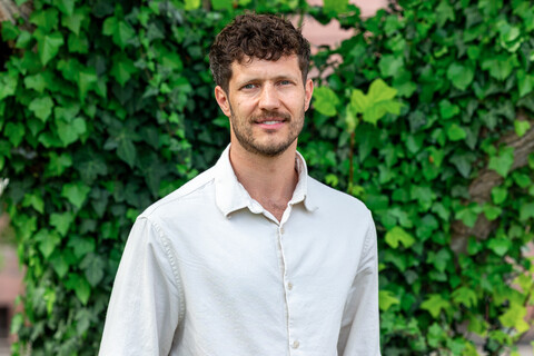 Porträt von Prof. Olbert. Er steht vor einer begrünten Wand, trägt ein weißes Hemd und lächelt in die Kamera.