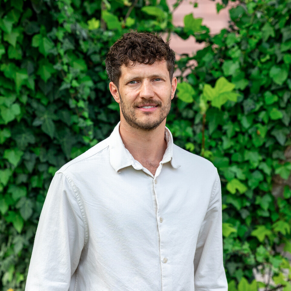 Porträt von Prof. Olbert. Er steht vor einer begrünten Wand, trägt ein weißes Hemd und lächelt in die Kamera.