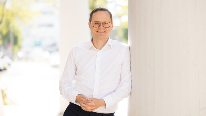 Eine Person trägt ein weißes Hemd sowie eine schwarze Hose und steht angelehnt an eine Säule. Die Person heißt Markus Strohmaier.