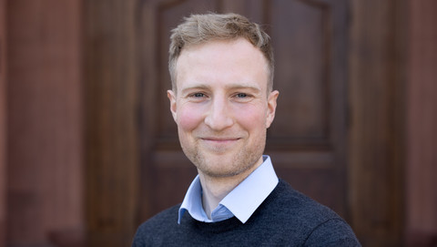 Felix Köhler. He is wearing grey sweater over a white shirt and is standing in front of a brown door.