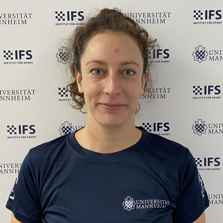 Francesca Bläse hat mittellanges, brunettes Haare. Sie trägt eine Brille und ein blaues T-Shirt mit der Aufschrift Universität Mannheim. Sie steht vor einer Stellwand, auf denen die Logos der Uni Mannheim und des IFS mehrfach abgebildet sind.