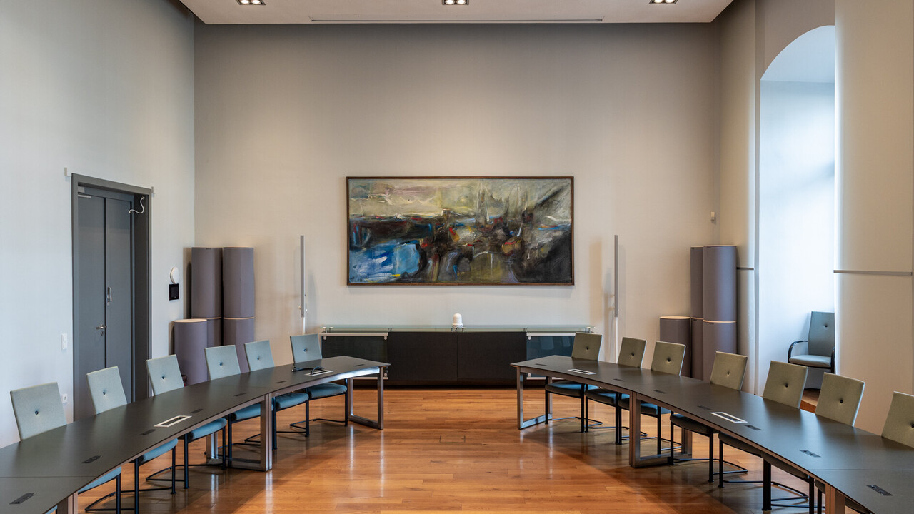 In einem Saal hängt ein großformatiges abstraktes Gemälde in Braun-, Blau und Grautönen und Streifen roter Farbe.