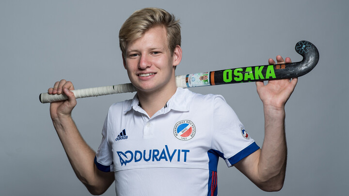 Linus Müller steht lächelnd vor einer grauen Wand. Er trägt ein weißes Trikot. Auf seinen Schultern liegt ein Hockeyschläger, den er mit zwei Händen hinter dem Kopf hält.