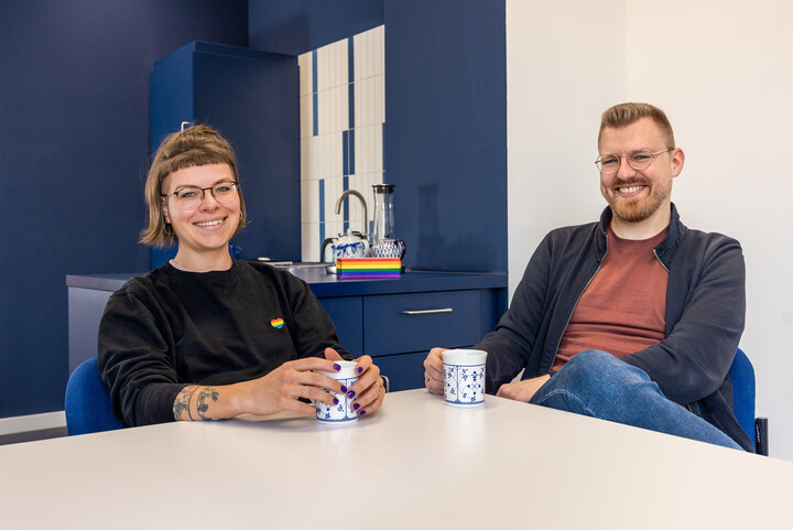 Janina Walker-Emig und Marcel Hörner sitzen an einem Tisch. Sie haben beide Kaffeetassen in den Händen.