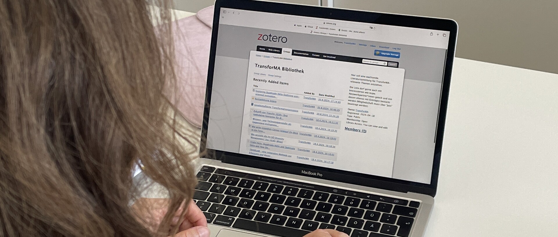 Eine Person mit schulterlangem braunem Haar sitzt an einem weißen Tisch und arbeitet an einem Laptop. Auf dem Bildschirm ist Zotero geöffnet, konkret eine Übersicht einer Bibliothek mit dem Titel ‚TransforMA Bibliothek‘. Im Hintergrund steht ein abschließbarer Aktenschrank.