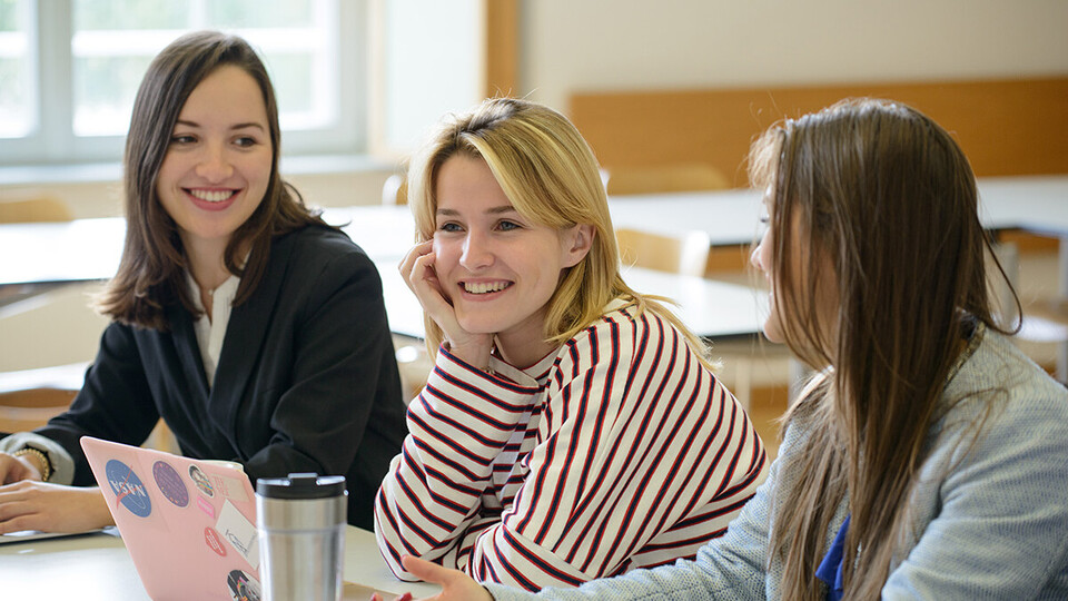 Drei Studierende sitzen lächelnd in Seminarraum