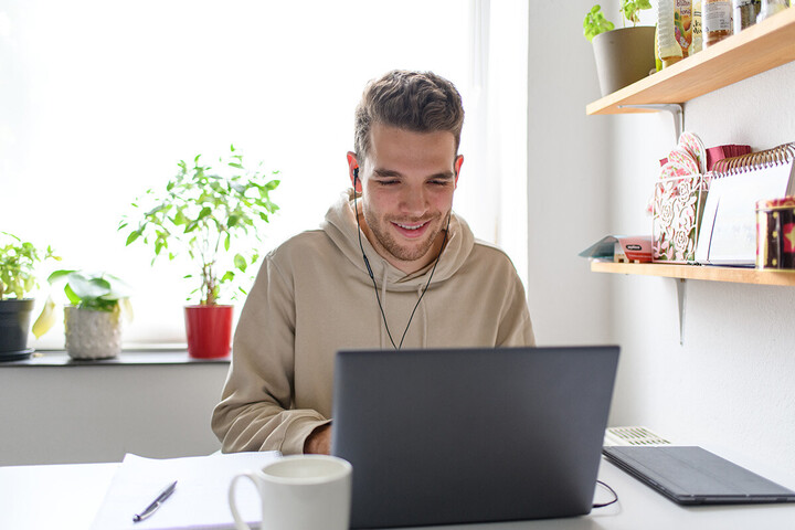 Ein Mann sitzt in einer Küche, hat Kopfhörer in den Ohren und tippt in seinen Laptop. Hinter ihm stehen Pflanzen, neben ihm hängt ein Regal.