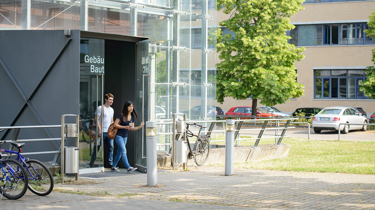 Ein Student und eine Studentin verlassen ein modernes, gläsernes Universitätsgebäude.