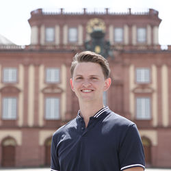 Sebastian Junker hat kurze braune Haar.e Er trägt ein blaues Polo-Hemd und steht vor dem Schloss.