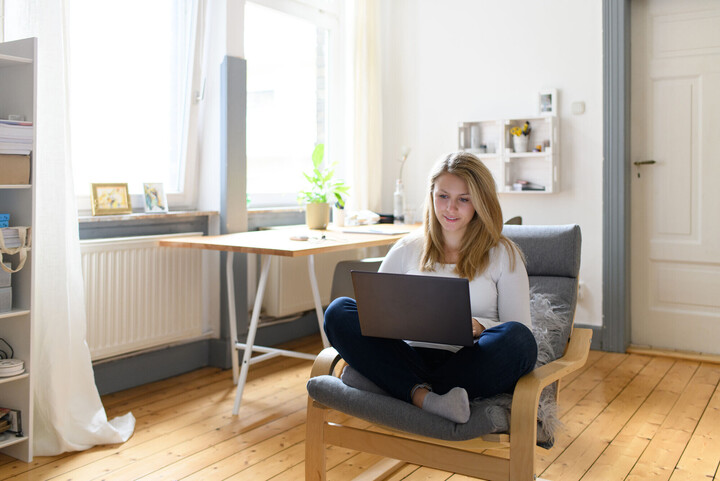 Eine Studentin sitzt in einem Sessel in ihrer Wohnung. Sie hat blonde Haare und hat einen Laptop auf dem Schoß. 