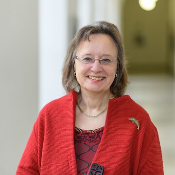 Portrait von Rosemarie Tracy. Sie trägt eine rote Bluse und einen roten Mantel und steht lächelnd in einem Gang.