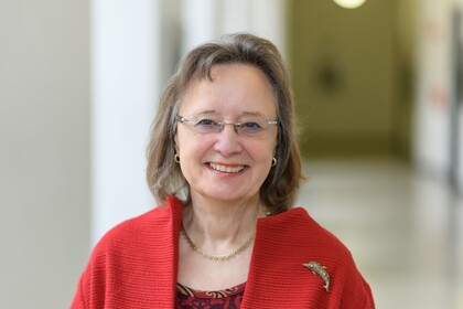 Portrait von Tracy Rosemary. Sie trägt eine rote Bluse und einen roten Mantel und steht lächelnd in einem Gang.