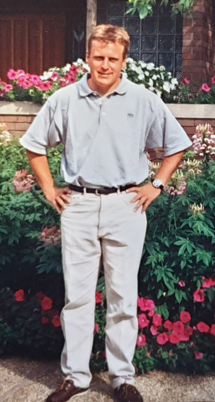 Stefan Fuchs trägt ein locker sitzendes graues Poloshirt und eine helle Hose und steht mit den Armen in die Hüfte gestemmt vor einem Blumenbeet