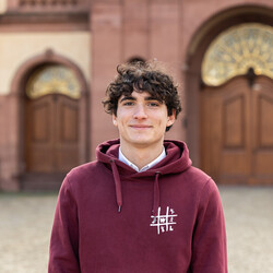 Emanuele Häge trägt einen weinroten Pullover mit dem Logo der Fachschaft VWL und lächelt freundlich in die Kamera.