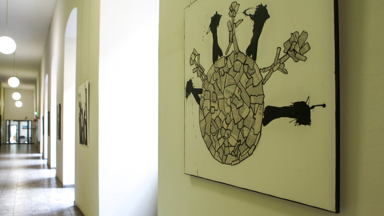 Ein Flur mit Bögen, welche an der rechten Wand entlanglaufen, ist geschmückt mit Kunstwerken, bestehend aus Zeitungscollagen und Farbklecks-Figuren.