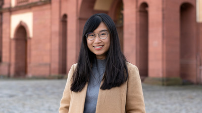 Eine lächelnde Person trägt einen grauen Rollkragenpullover sowie einen hellbraunen Mantel und steht vor dem Schloss der Uni Mannheim. Die Person heißt Yu-Han Mao.