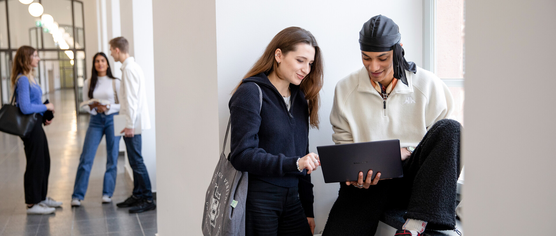Zwei Studierende schauen auf einen Laptop, während der Student auf einer Fensterbank sitzt und die Studentin neben ihm steht. Im Hintergrund sind weitere Studierende zu sehen, die sich unterhalten.