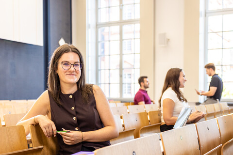 Eine Studentin sitzt in einer Bankreihe im Hörsaal. Sie schaut direkt in die Kamera und lächelt. Im Hintergrund sitzen weitere Studierende.