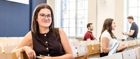 Eine Studentin sitzt in einer Bankreihe im Hörsaal. Sie schaut direkt in die Kamera und lächelt. Im Hintergrund sitzen weitere Studierende.