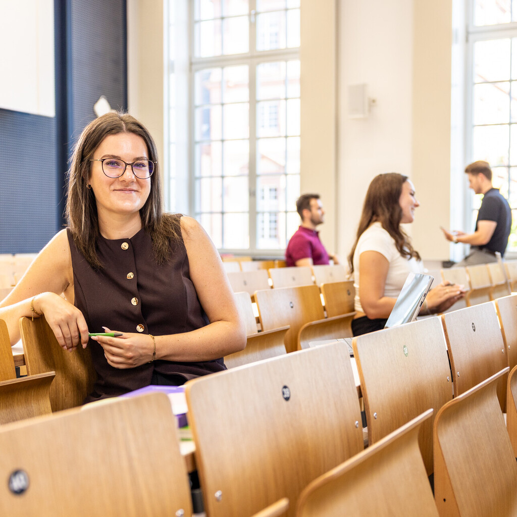 Eine Studentin sitzt in einer Bankreihe im Hörsaal. Sie schaut direkt in die Kamera und lächelt. Im Hintergrund sitzen weitere Studierende.
