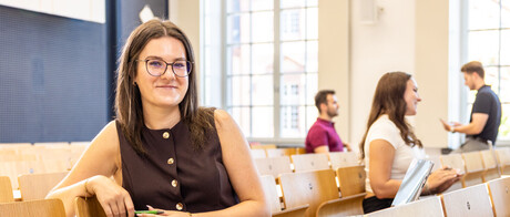 Eine Studentin sitzt in einer Bankreihe im Hörsaal. Sie schaut direkt in die Kamera und lächelt. Im Hintergrund sitzen weitere Studierende.