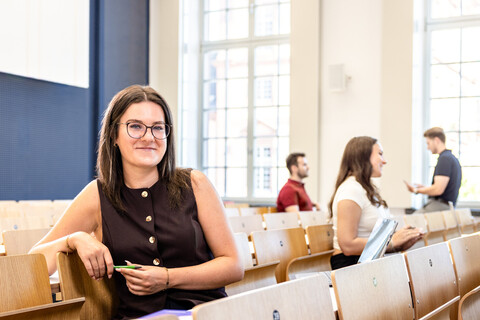 Eine Studentin sitzt in einer Bankreihe im Hörsaal. Sie schaut direkt in die Kamera und lächelt. Im Hintergrund sitzen weitere Studierende.