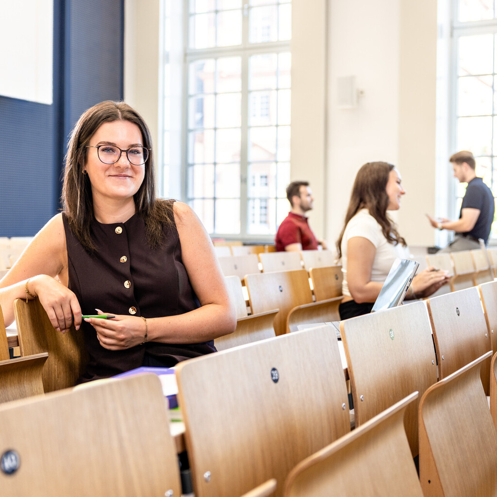 Eine Studentin sitzt in einer Bankreihe im Hörsaal. Sie schaut direkt in die Kamera und lächelt. Im Hintergrund sitzen weitere Studierende.