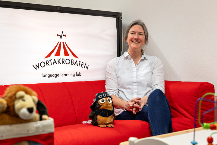 Nicole ALtvater-Mackensen sitzt auf einer roten Couch neben einem Stofftier. Hinter ihr ist das Logo der Wortakrobaten zu sehen.