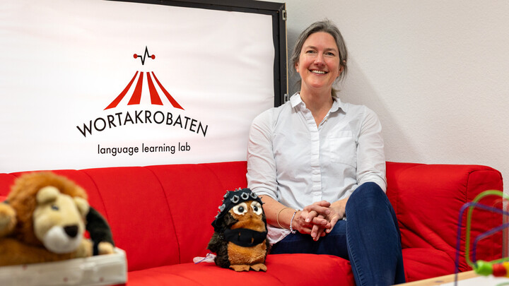 Nicole ALtvater-Mackensen sitzt auf einer roten Couch neben einem Stofftier. Hinter ihr ist das Logo der Wortakrobaten zu sehen.
