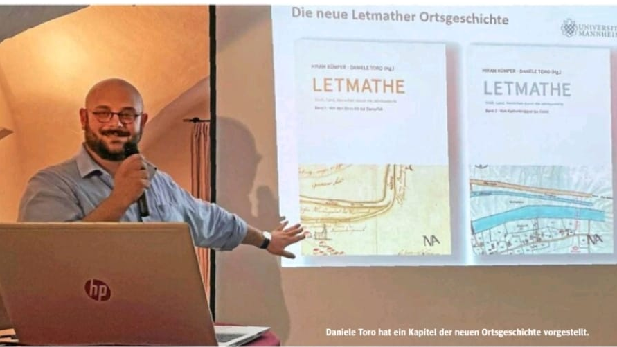 Zeitungsausschnitt mit einem männlichen Vortragenden vor der Leinwand, die ein Buchcover zeigt