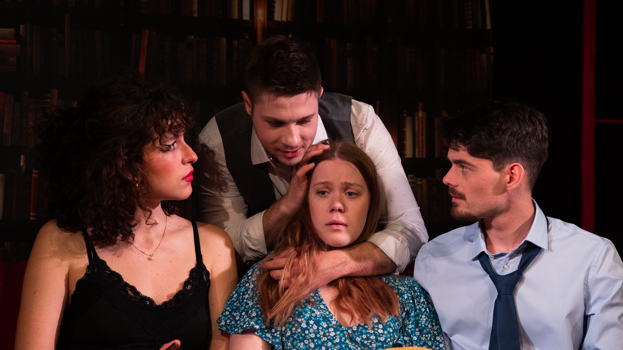 Szenenbild des Unitheaters. Ein Student hält den Kopf einer vor sich sitzenden Studentin fest und zwei weitere Studierende schauen zu.