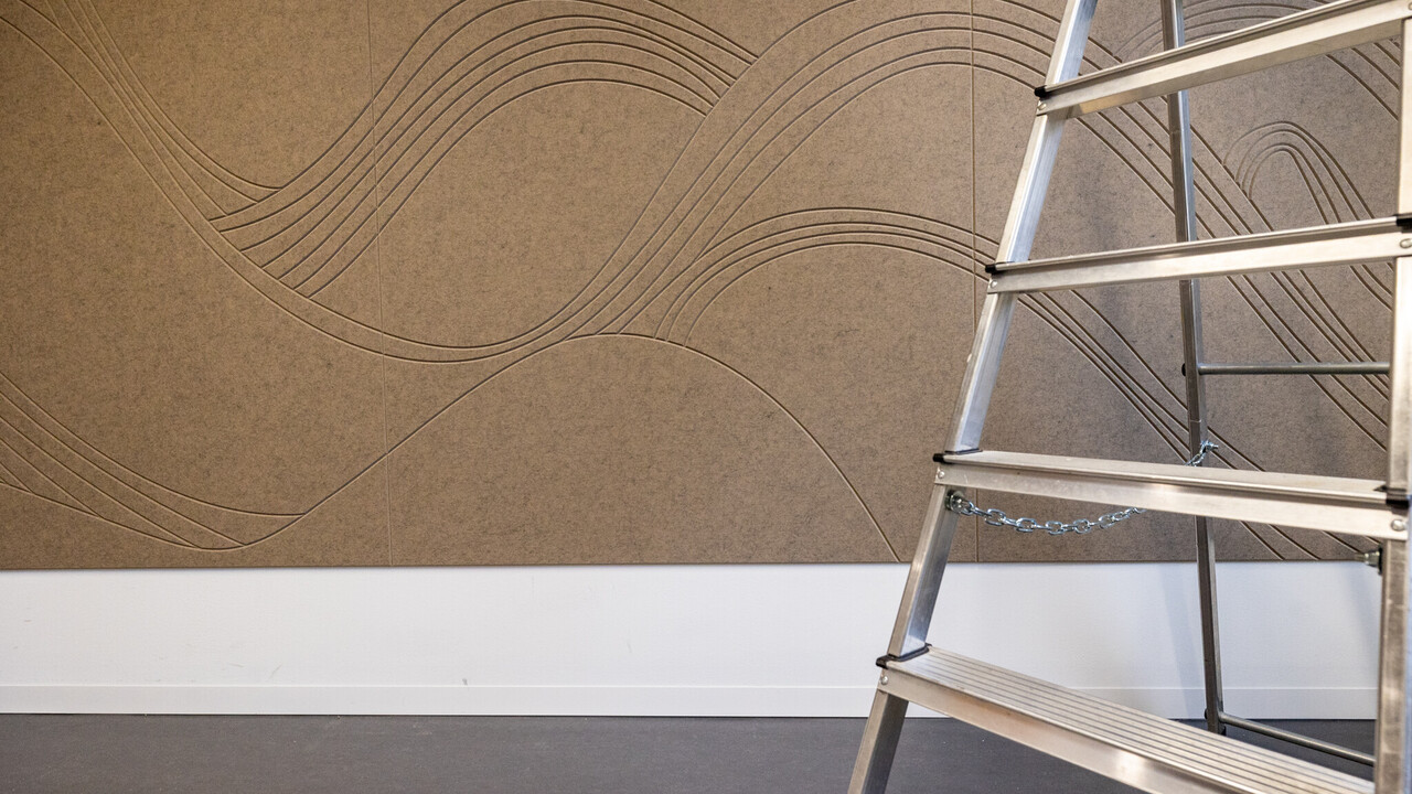 Zu sehen ist ein Wandausschnitt mit einer braunen, schallhemmenden Filzwand, die mit Wellen dekoriert ist und der Teil einer Leiter