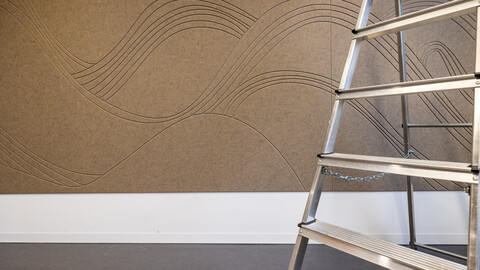 Zu sehen ist ein Wandausschnitt mit einer braunen, schallhemmenden Filzwand, die mit Wellen dekoriert ist und der Teil einer Leiter