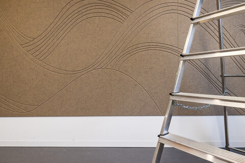 Zu sehen ist ein Wandausschnitt mit einer braunen, schallhemmenden Filzwand, die mit Wellen dekoriert ist und der Teil einer Leiter