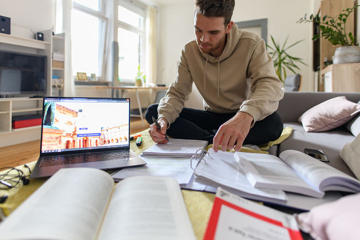 Student sitzt auf dem Boden eines Zimmers und hat viele Bücher, Skripte und einen Laptop vor sich. Er lernt. 