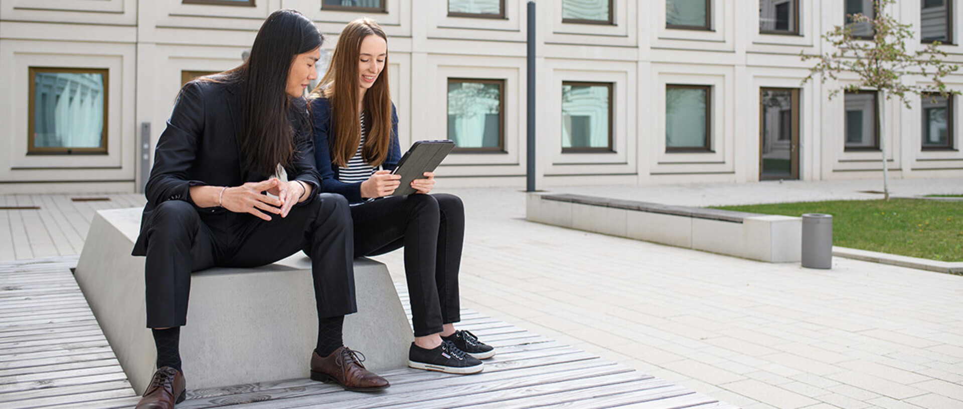 A man and a woman are sitting in the courtyard of the B6, 30-32 building and reading the news on a tablet.