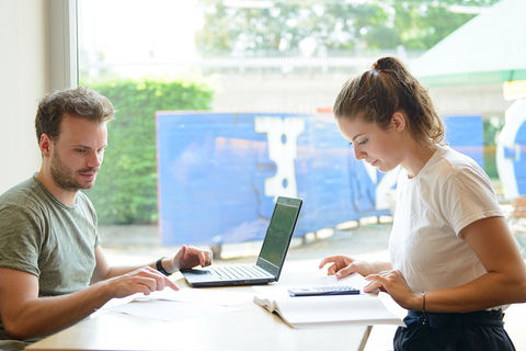 Eine Studentin und ein Student arbeiten gemeinsam an einem Tisch. Vor ihnen liegen ein Laptop, ein Taschenrechner und ein Buch.