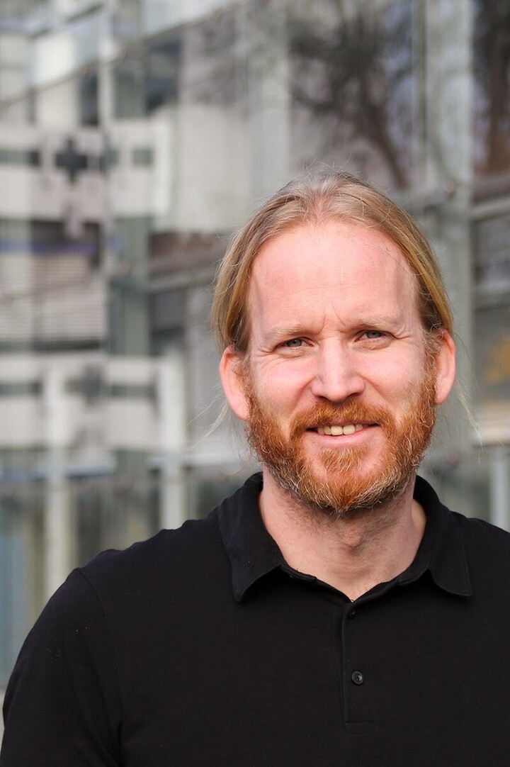 Portrait eines Mannes mit schwarzem Hemd, der vor dem B6-Gebäude steht. 
