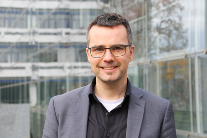 Portrait von Marc Debus. Er trägt einen grauen Blazer und ein schwarzes Hemd.