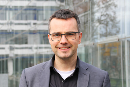 Portrait von Marc Debus. Er trägt einen grauen Blazer und ein schwarzes Hemd.