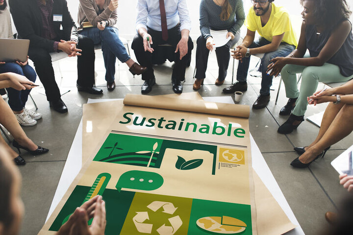 Personen sitzen im Stuhlkreis um ein auf dem Boden liegendes Plakat herum. Auf diesem steht Sustainable und mehrere Symbole.