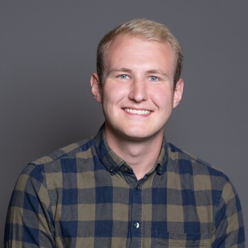 Tobias Kreuz. Er trägt ein blau-grün kariertes Hemd und steht vor einer dunklen Wand.