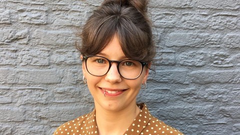 Lena Arnold. She is wearing a khaki-colored blouse and is leaning against a grey stonewall.